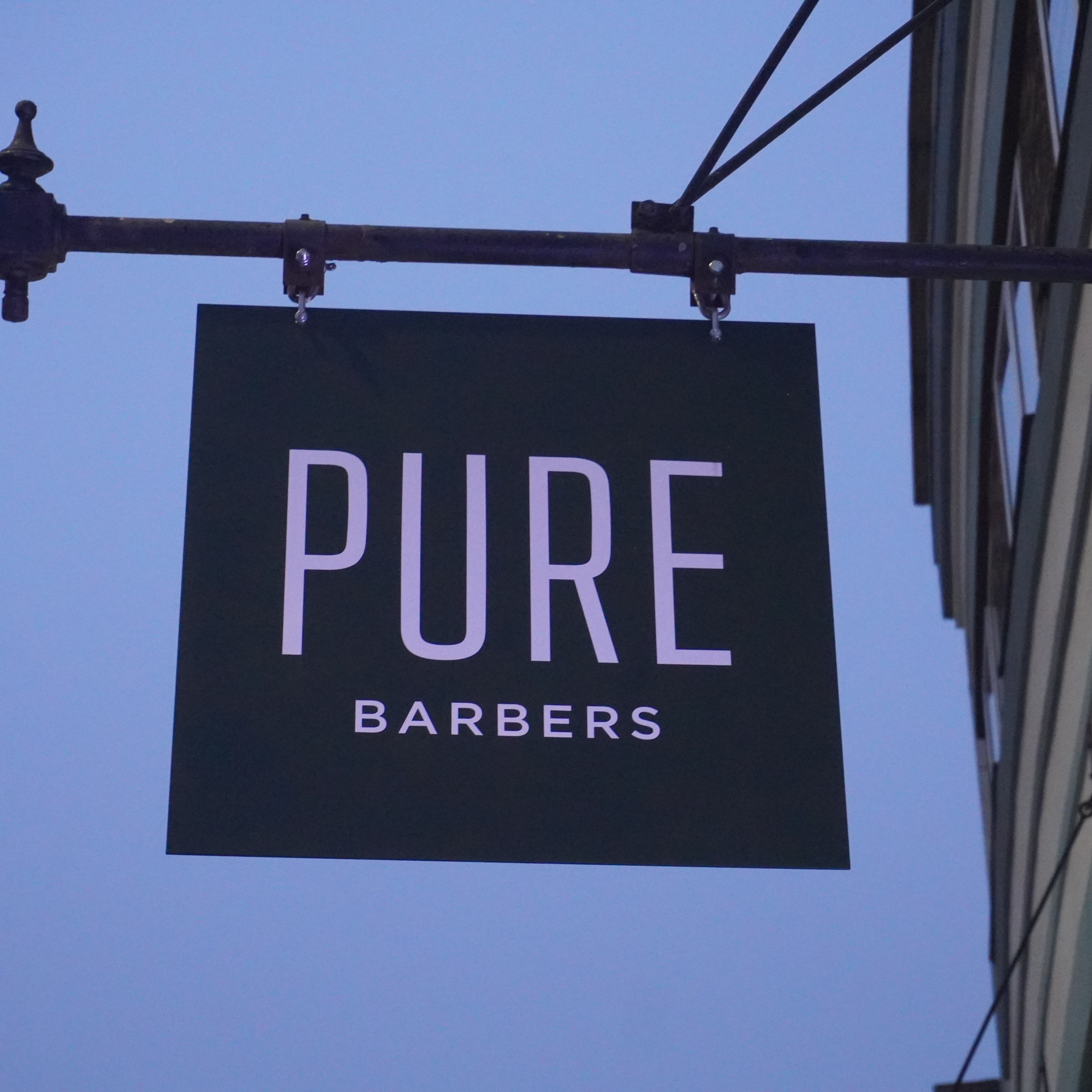 Signage of Pure Barbers hanging outdoors with evening sky in the background.